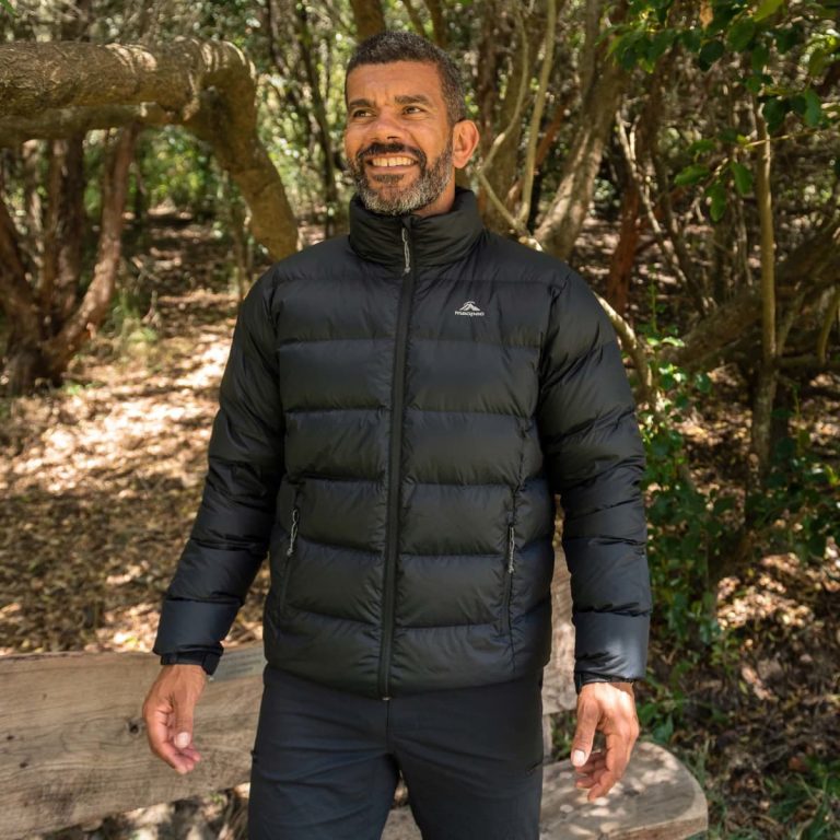 A man wearing a black puffer jacket stands outdoors, surrounded by trees and foliage. The jacket features a logo.