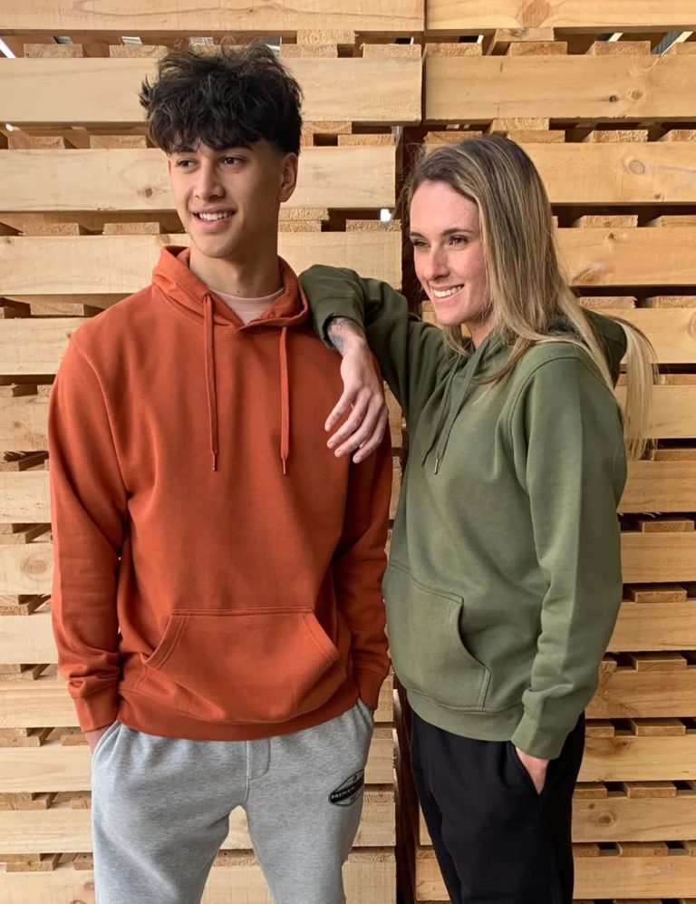 A brown hoodie and a green hoodie are worn by two models against a wooden backdrop. Both hoodies have a front pocket.